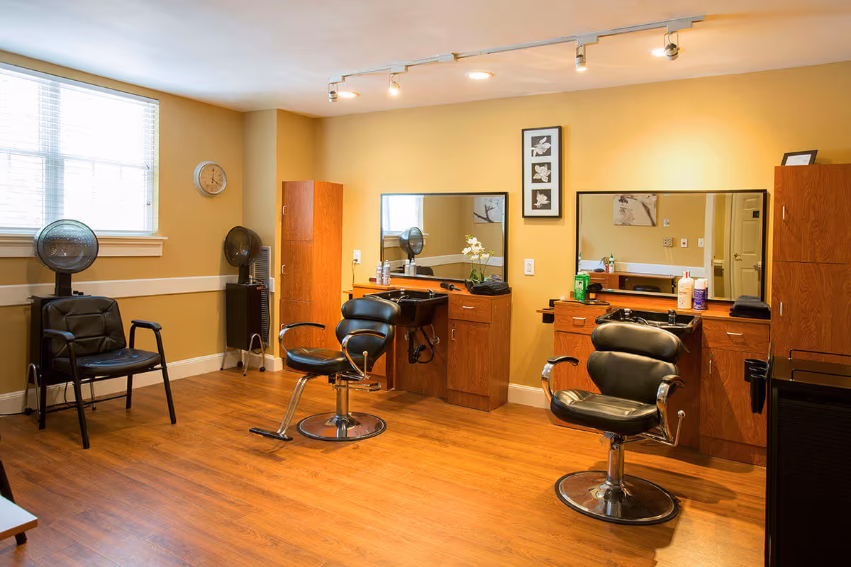 Interior of a hair salon room with two black salon chairs in front of wooden cabinets and mirrors. There are two hair dryers on stands near a window with blinds, a clock on the wall, and various hair care products on the counters. The room has wooden flooring and yellow walls.