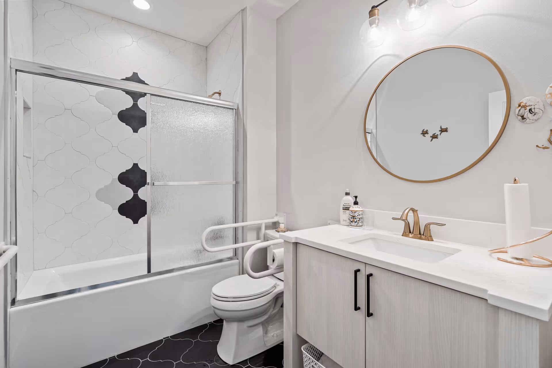 A modern bathroom featuring a bathtub with sliding glass doors and decorative white and black tiles on the wall. There is a toilet with safety grab bars installed on both sides. A vanity with a white countertop, gold faucet, and a round mirror above it is visible. The bathroom has a clean and bright appearance with white walls and black floor tiles.
