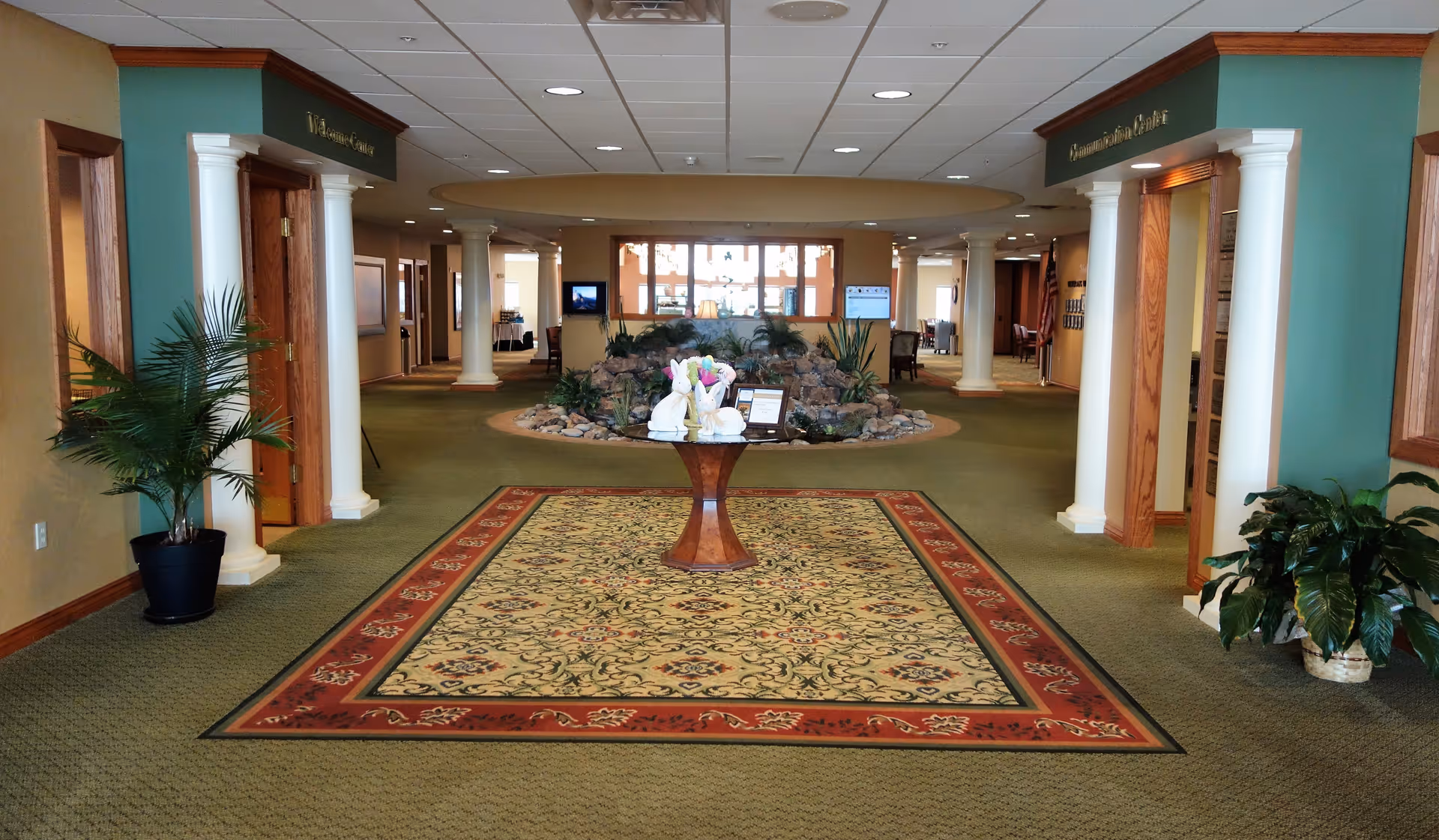 Interior view of a retirement community hallway with green carpet and a decorative rug in the center. A round wooden table with floral and decorative items is placed on the rug. The hallway features white columns, potted plants, and a rock garden with greenery in the background. Doors and seating areas are visible along the sides.
