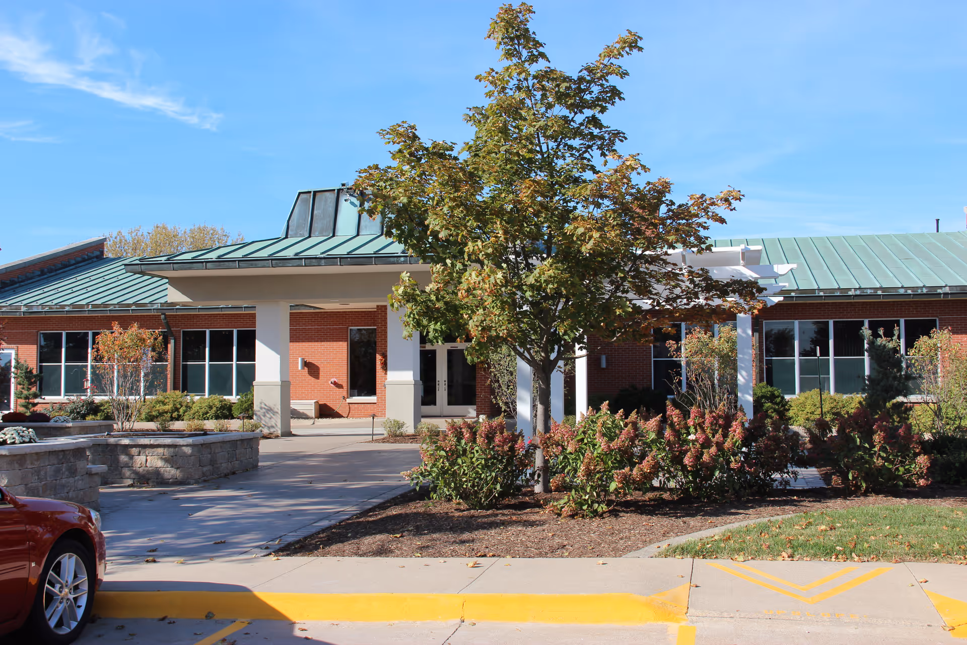Exterior view of The Alverno Senior Care Community building with a green metal roof, brick walls, large windows, and a covered entrance. There is a tree and landscaped bushes in front of the entrance, a sidewalk, and a red car partially visible on the left side.
