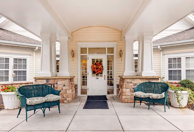 Entrance to a senior living facility with a white door decorated with an autumn wreath, flanked by two stone pillars and two green wicker benches with cushions on either side. Large white planters with flowers are placed near the benches.