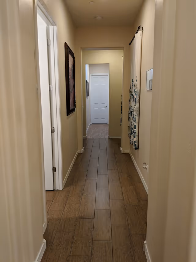 A narrow hallway with wood-look tile flooring and cream-colored walls. There are doorways on the left side and artwork hanging on the walls. A white door is visible at the end of the hallway.