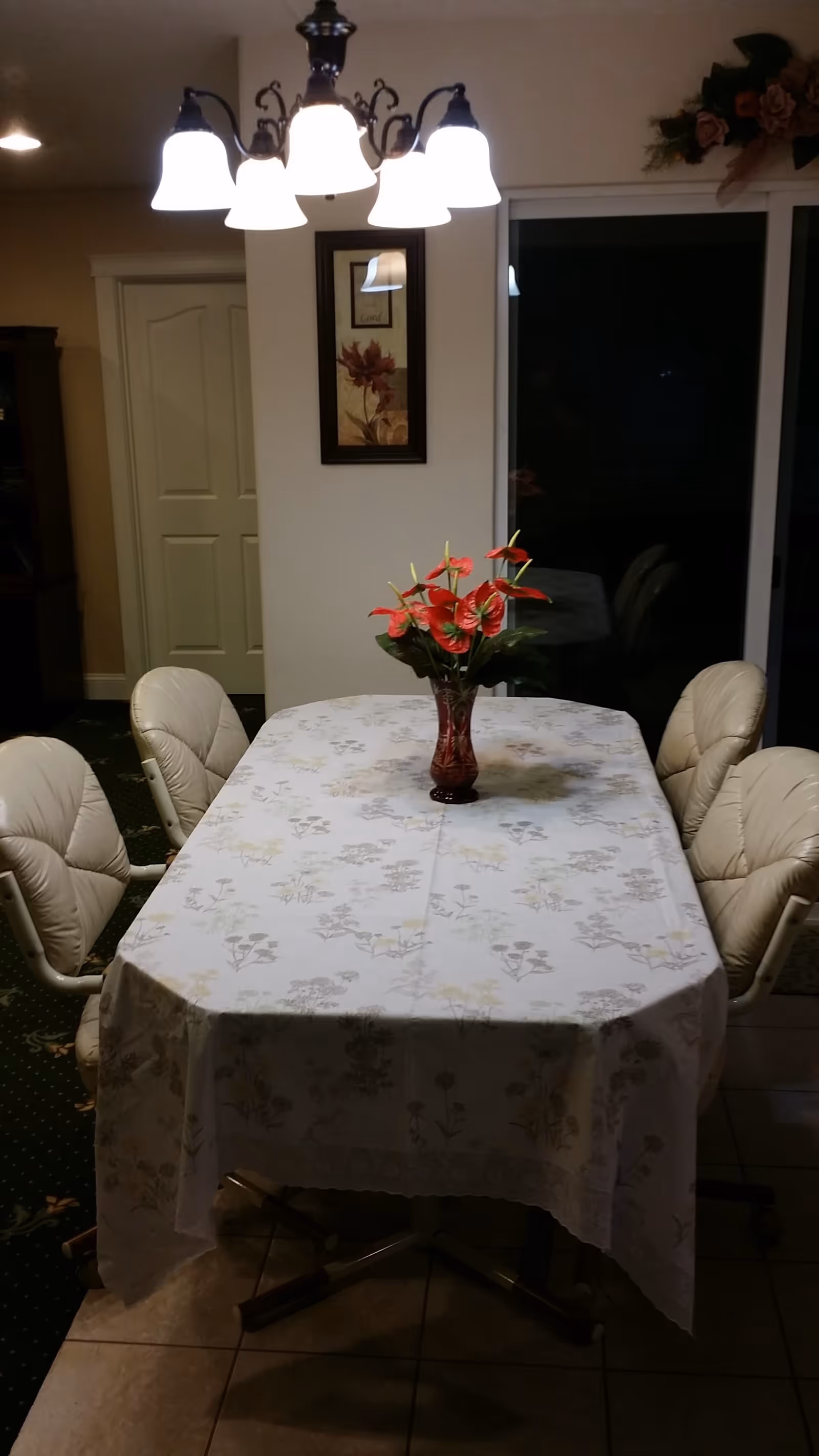 A dining room with a rectangular table covered with a floral tablecloth. There are six cushioned chairs around the table. A vase with red flowers is placed in the center of the table. Above the table hangs a chandelier with five lit lamps. A framed floral picture is on the wall behind the table, and a sliding glass door is visible to the right.