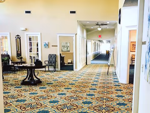 Interior view of a senior living facility hallway with patterned carpet, beige walls, and several doorways leading to rooms. There is a round table with chairs on the left side and a long corridor extending into the distance with ceiling lights and an exit sign.