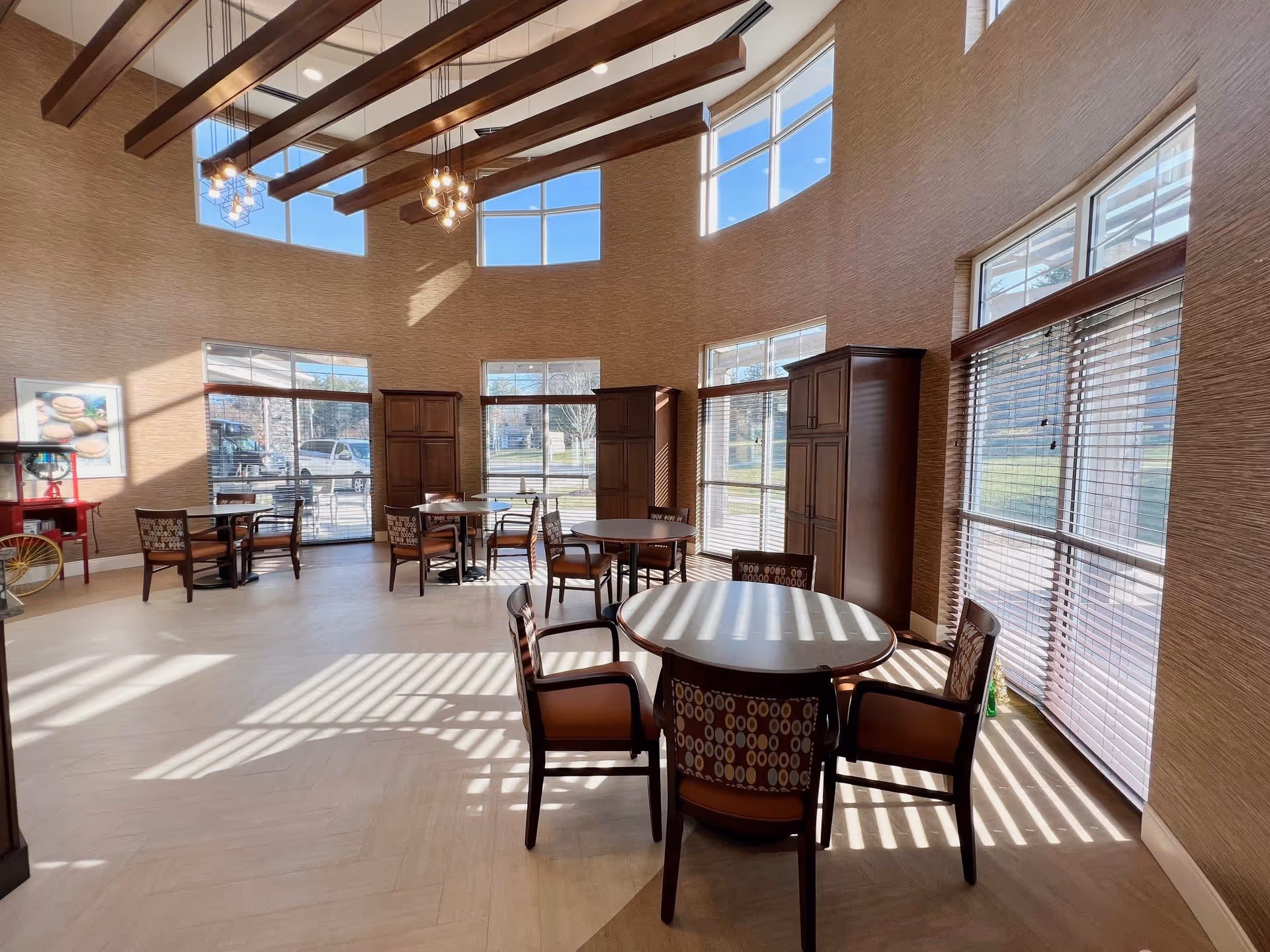 A bright, spacious dining area with round tables and chairs arranged neatly. The room features large windows with blinds allowing natural light to cast shadows on the floor. The walls are decorated with a textured finish and a framed picture. The ceiling has exposed wooden beams with modern hanging light fixtures.