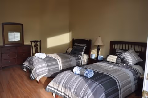 A bedroom with two single beds featuring gray and black plaid bedding, each with a rolled towel placed on top. There is a wooden dresser with a mirror and a wooden chair against the wall. A bedside table with a lamp and a box of tissues is positioned between the beds. Sunlight streams through a window, casting shadows on the beds and wall.