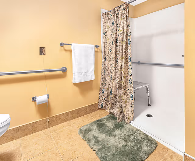 Accessible bathroom with beige tile floor and walls, grab bars, a towel and toilet paper holder, a walk-in shower with patterned curtain and shower chair, and a green bath mat.