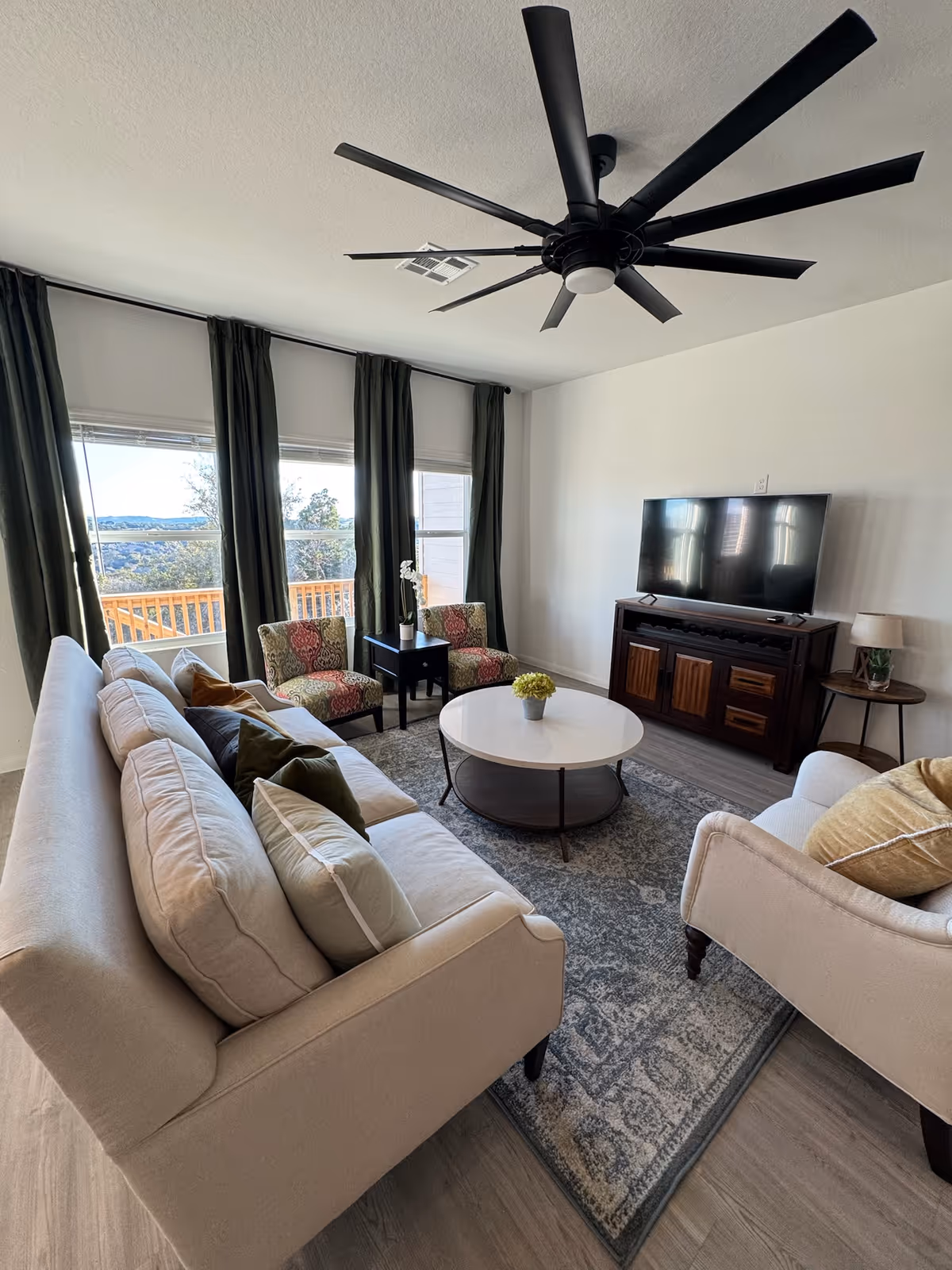 A bright living room with a large beige sofa adorned with multiple cushions, two patterned armchairs, and a round white coffee table with a small plant on it. The room has large windows with dark curtains, a ceiling fan with multiple blades, a TV on a wooden stand, and a side table with a lamp. The floor is covered with a patterned rug.
