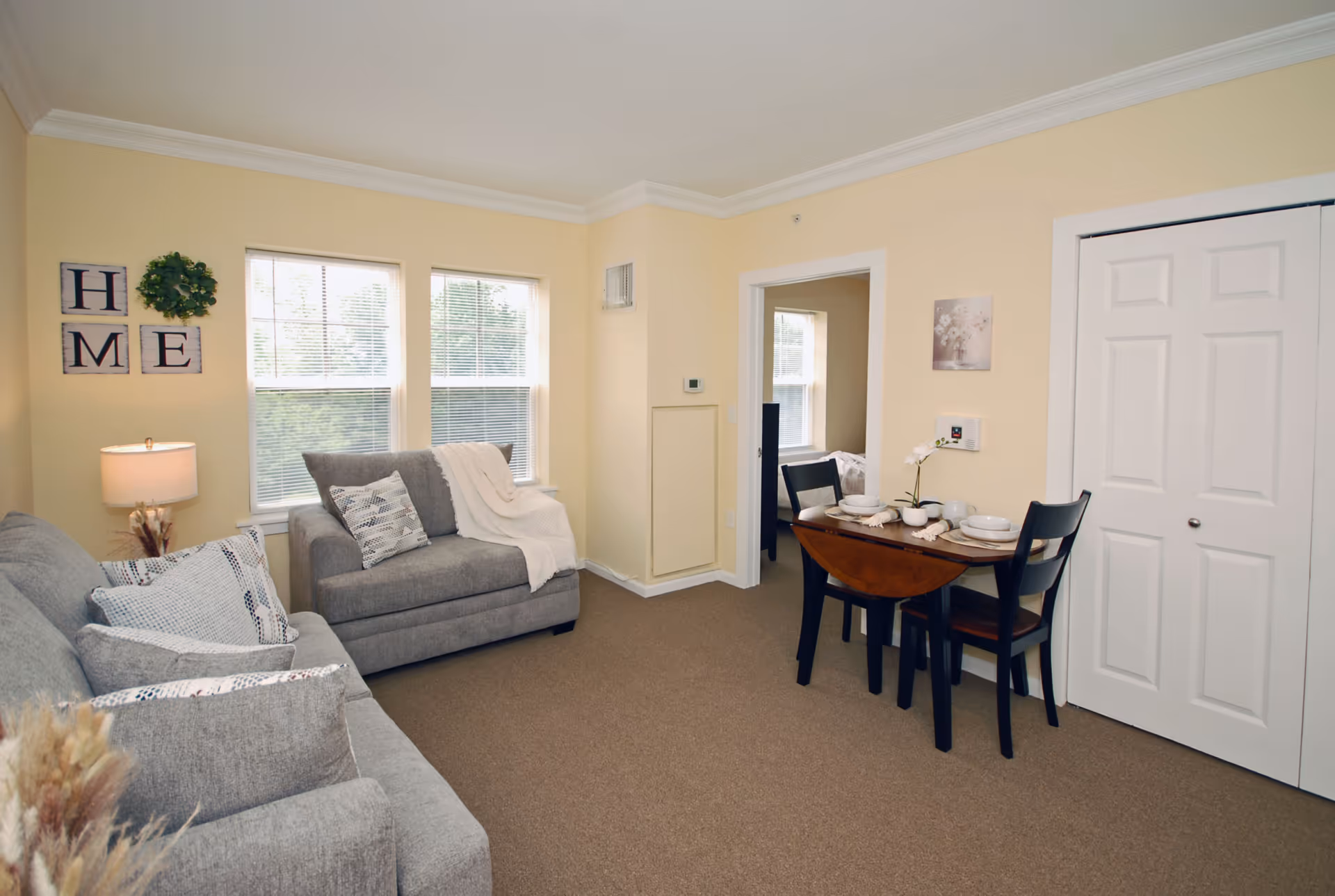 A cozy living room with beige walls and carpeted floor featuring two gray sofas with patterned pillows and a white throw blanket. There is a small wooden dining table set for two with black chairs near a white closet door. Two windows with blinds allow natural light into the room. Wall decor includes a wreath and framed letters spelling 'HOME'.