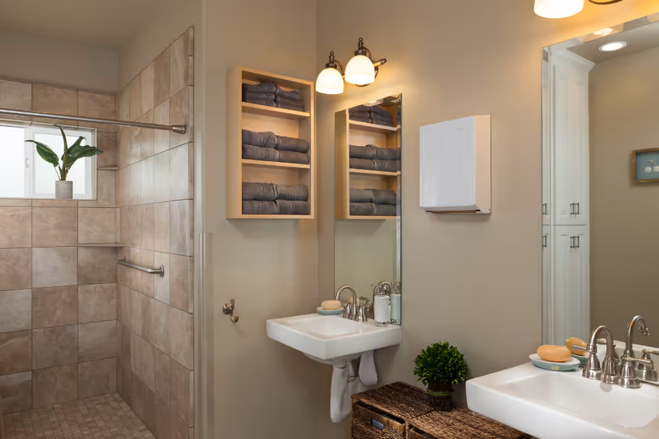 A clean and modern bathroom with a tiled walk-in shower on the left, a small window with a potted plant on the windowsill, a wall-mounted sink with soap and lotion dispensers, a large mirror, a wall-mounted cabinet filled with neatly folded dark towels, and a wicker basket with a small green plant on top.