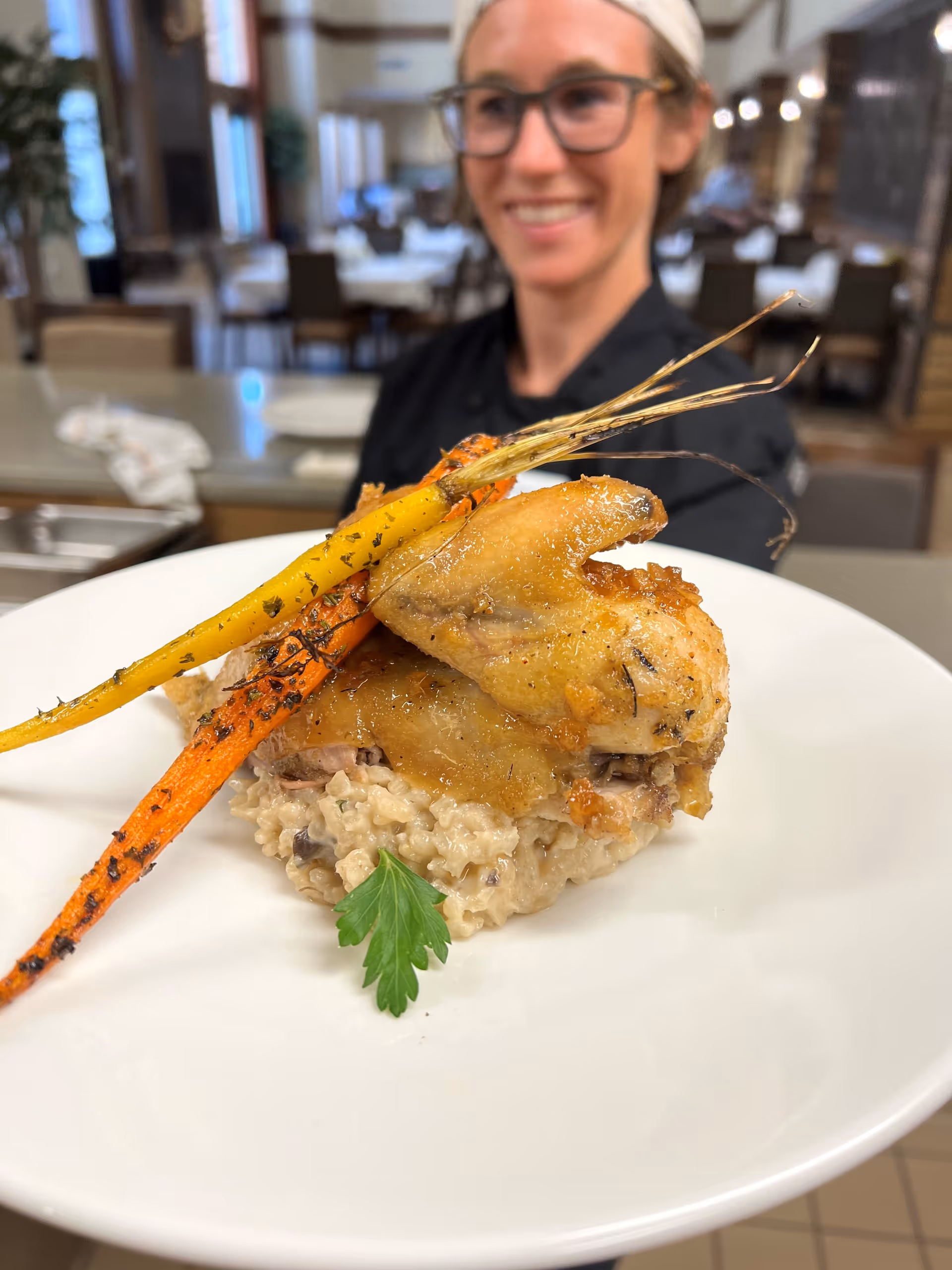 A close-up of a plated dish featuring roasted chicken wings on a bed of creamy risotto, garnished with two roasted carrots and a sprig of parsley. In the background, a smiling chef wearing glasses and a headband is slightly out of focus in a dining area with tables and chairs.