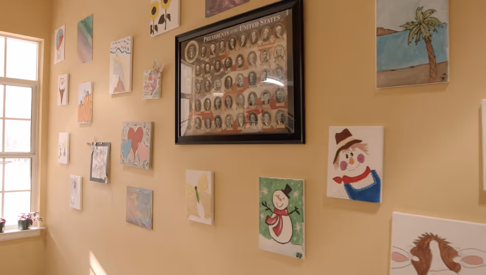 A beige interior wall decorated with small colorful paintings and a framed 'Presidents of the United States' poster beside a window.