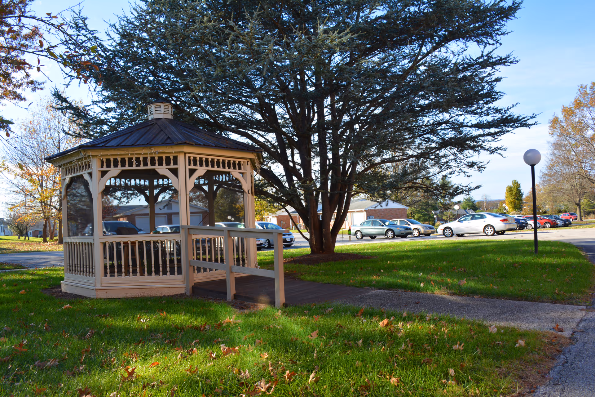 A white wooden gazebo with a dark roof situated on a grassy area with a paved walkway leading to it. Behind the gazebo, there is a large tree and a parking lot with several parked cars. The sky is clear and blue.