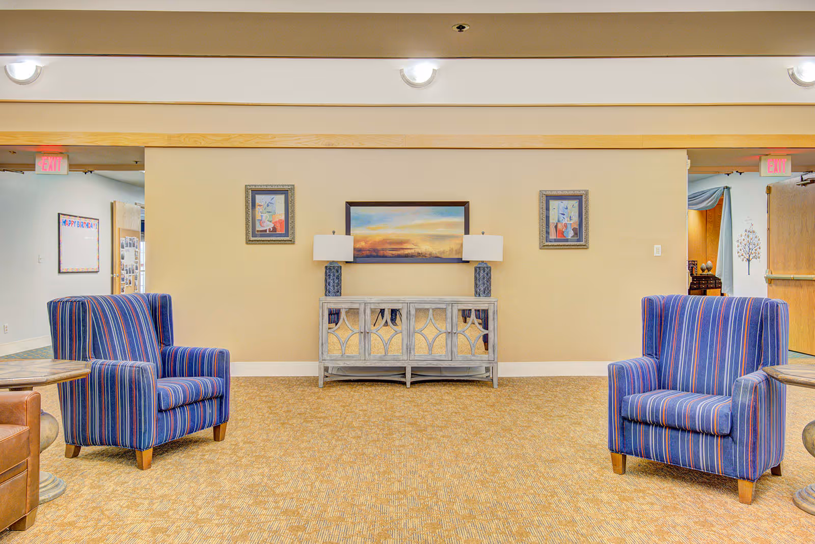 A sitting area in an assisted living facility with two blue striped armchairs facing each other, a wooden sideboard with mirrored doors against a beige wall, two table lamps with blue bases on the sideboard, and three framed artworks hanging above it. The carpet is patterned in beige tones, and there are exit signs visible in the background.