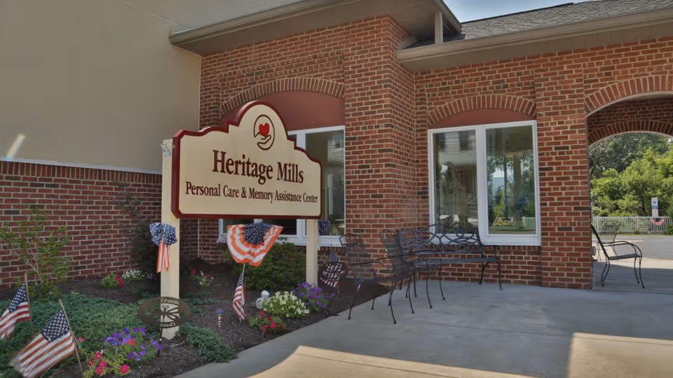 Entrance area of Heritage Mills Personal Care & Memory Assistance Center with a large sign displaying the facility name. The building has red brick walls, windows, and a covered walkway with metal benches. There are small American flags and patriotic decorations in the flower beds near the sign.