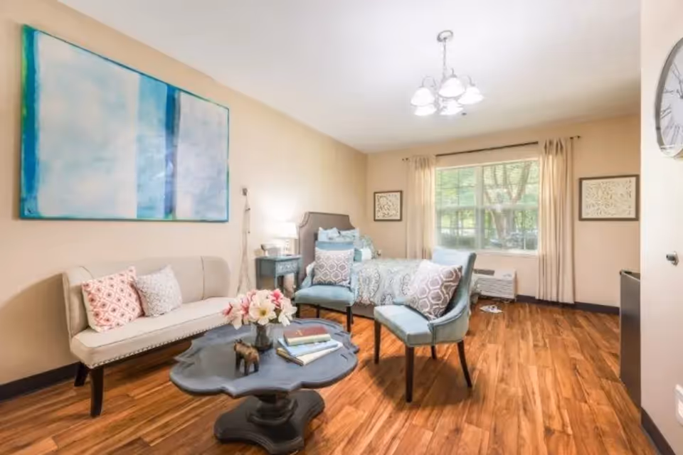 A cozy, well-lit bedroom with a bed, two blue chairs with patterned pillows, a beige loveseat with pillows, a dark wooden coffee table with books and flowers, a large window with beige curtains, and a large abstract painting on the wall.