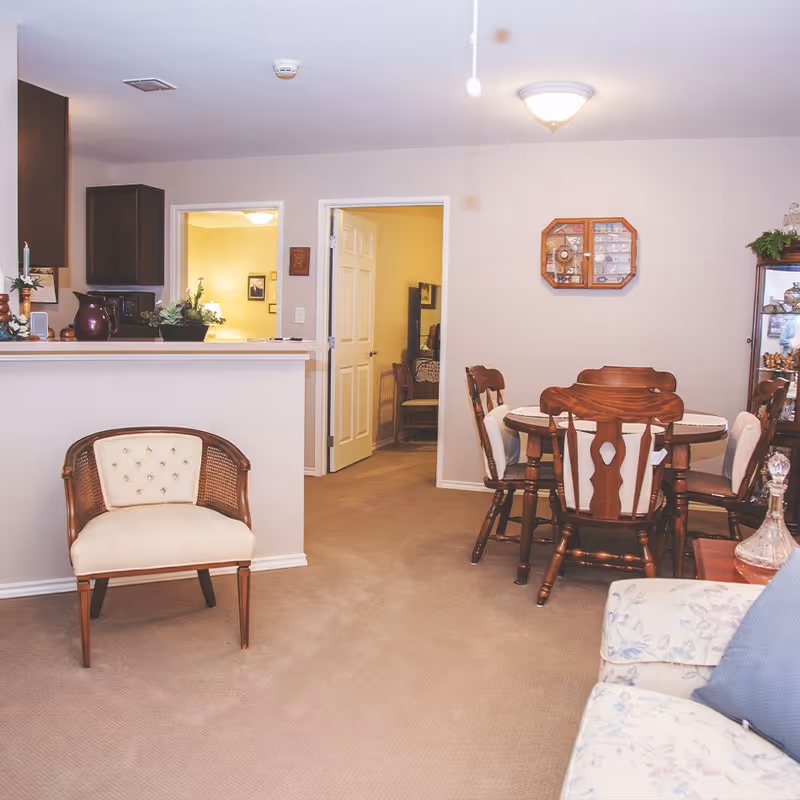 Open-plan interior showing a dining table with wooden chairs, an upholstered armchair, and a view into adjoining rooms.