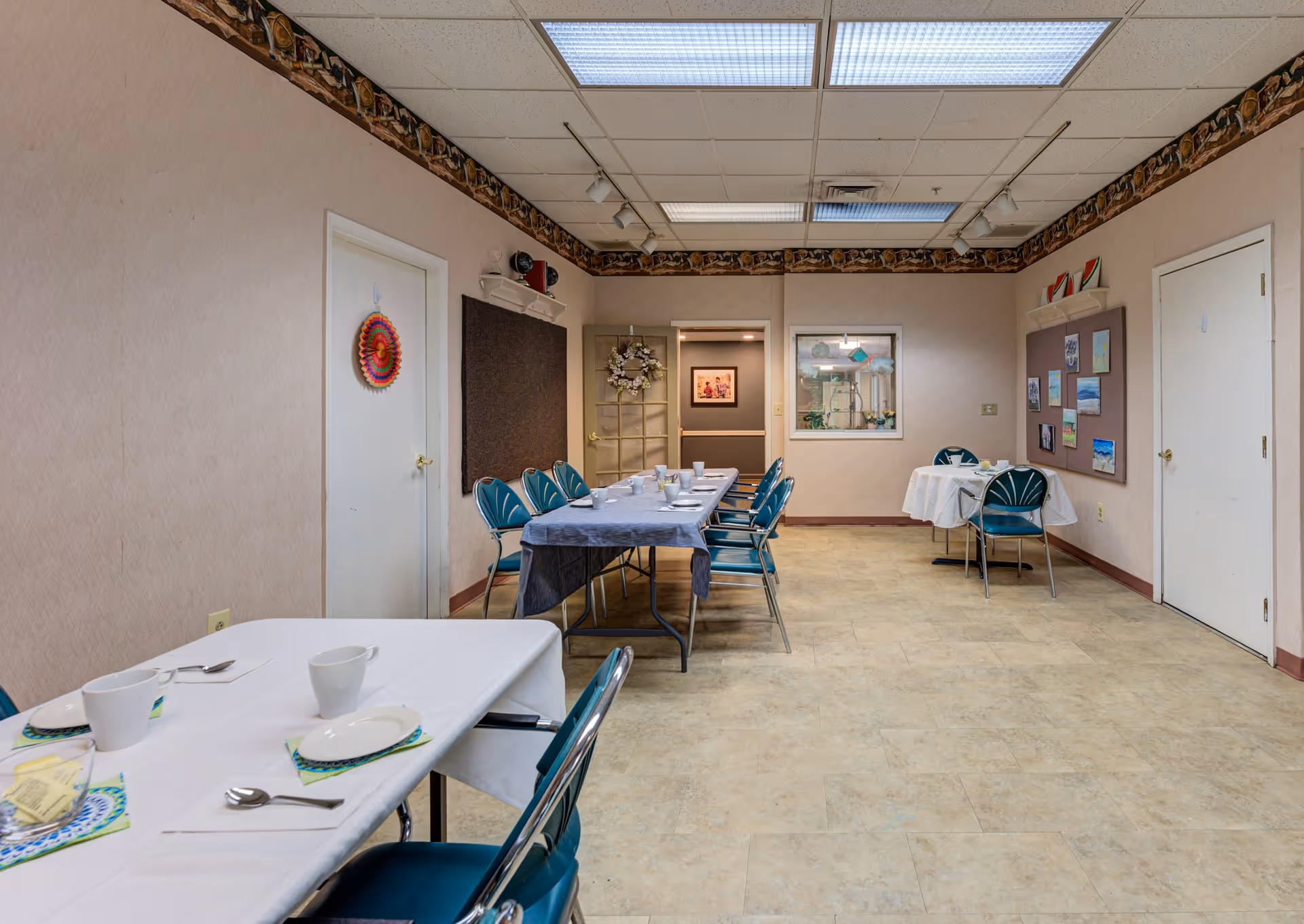 A dining room with several tables covered with white and blue tablecloths, set with cups, plates, and utensils. Blue chairs are arranged around the tables. The room has beige walls with a decorative border near the ceiling, fluorescent lighting, and bulletin boards with pictures and decorations on the walls.