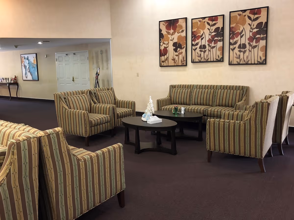 A seating area in a senior living facility with striped upholstered armchairs and a matching sofa arranged around two black coffee tables. The tables hold a small white decorative tree and a tissue box. The walls are beige with three framed floral artworks hanging above the sofa. In the background, there is a hallway with a white door and a console table with a painting above it.
