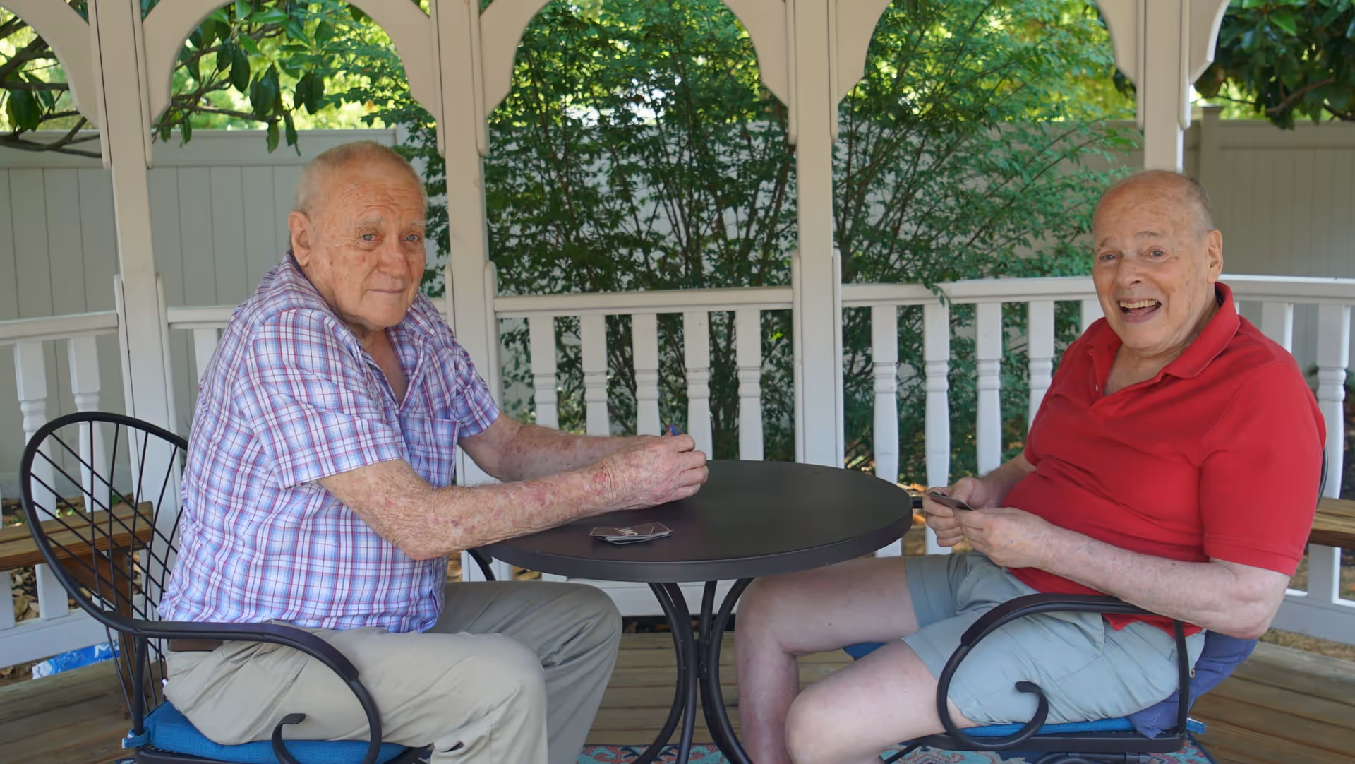 Two elderly men sitting at a round black metal table under a white gazebo, playing cards. One man is wearing a purple plaid shirt and beige pants, while the other is wearing a red polo shirt and light gray shorts. Green foliage is visible in the background.