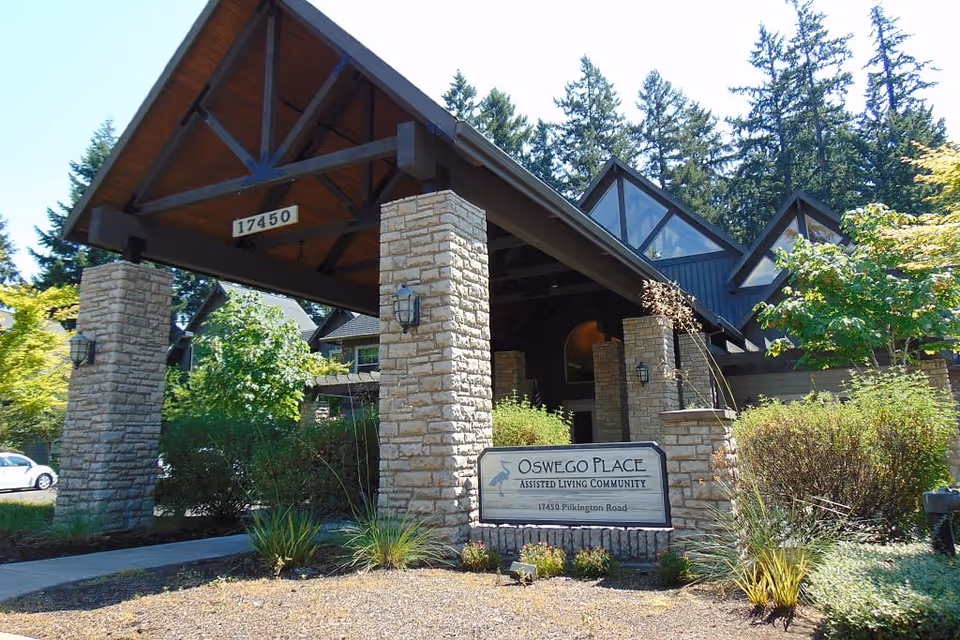 Entrance of Oswego Place Assisted Living Community featuring a large covered driveway supported by stone pillars, surrounded by greenery and trees under a clear sky.