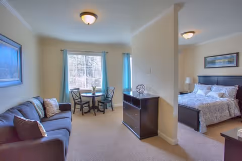 A cozy living space in an assisted living facility featuring a dark sofa with pillows on the left, a small round table with two chairs near a window with blue curtains, and a bedroom area partially visible on the right with a bed, nightstand, lamp, and framed artwork on the wall.