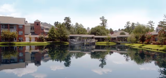 A serene outdoor view of a pond reflecting surrounding trees and buildings at The Forum at The Woodlands senior living facility under a clear blue sky.