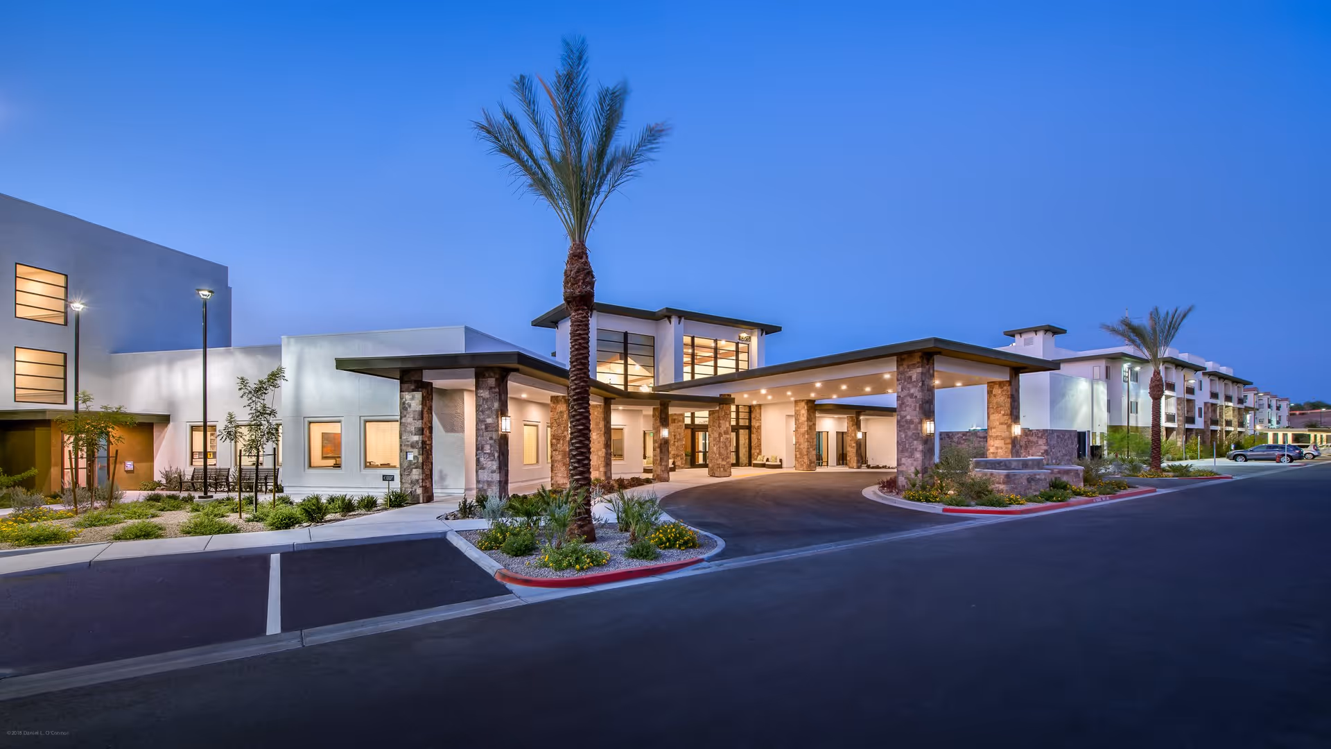 Exterior view of Revel Nevada senior living facility at dusk, showing a modern building with stone pillars, large windows, palm trees, and a covered entrance with driveway.