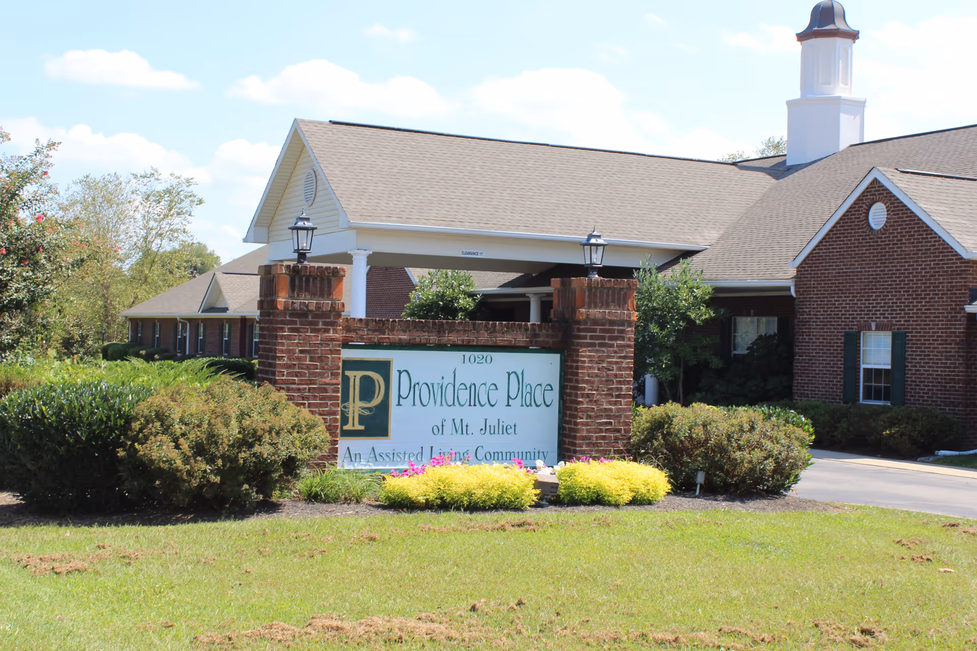 Exterior view of Providence Place of Mt. Juliet, an assisted living community, showing a brick sign with the facility name and a building with a covered entrance and cupola in the background surrounded by greenery and a lawn.