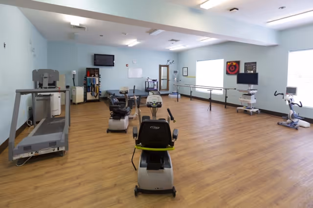 Rehabilitation exercise room with stationary and recumbent bikes, a treadmill, parallel bars and TVs in a light-blue, wood-floored space.
