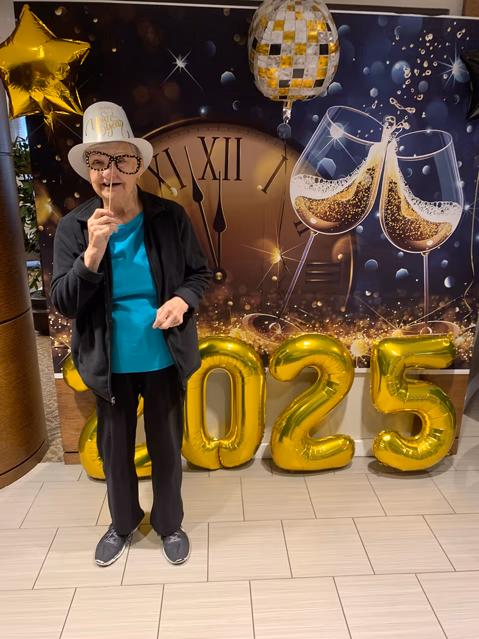 An elderly woman wearing a white 'Happy New Year' hat and holding a stick with glasses in front of her face, standing indoors on a tiled floor. Behind her is a festive backdrop featuring a large clock showing midnight, two clinking champagne glasses, and gold balloons forming the numbers '2025'. There are also star-shaped and disco ball balloons as part of the decoration.