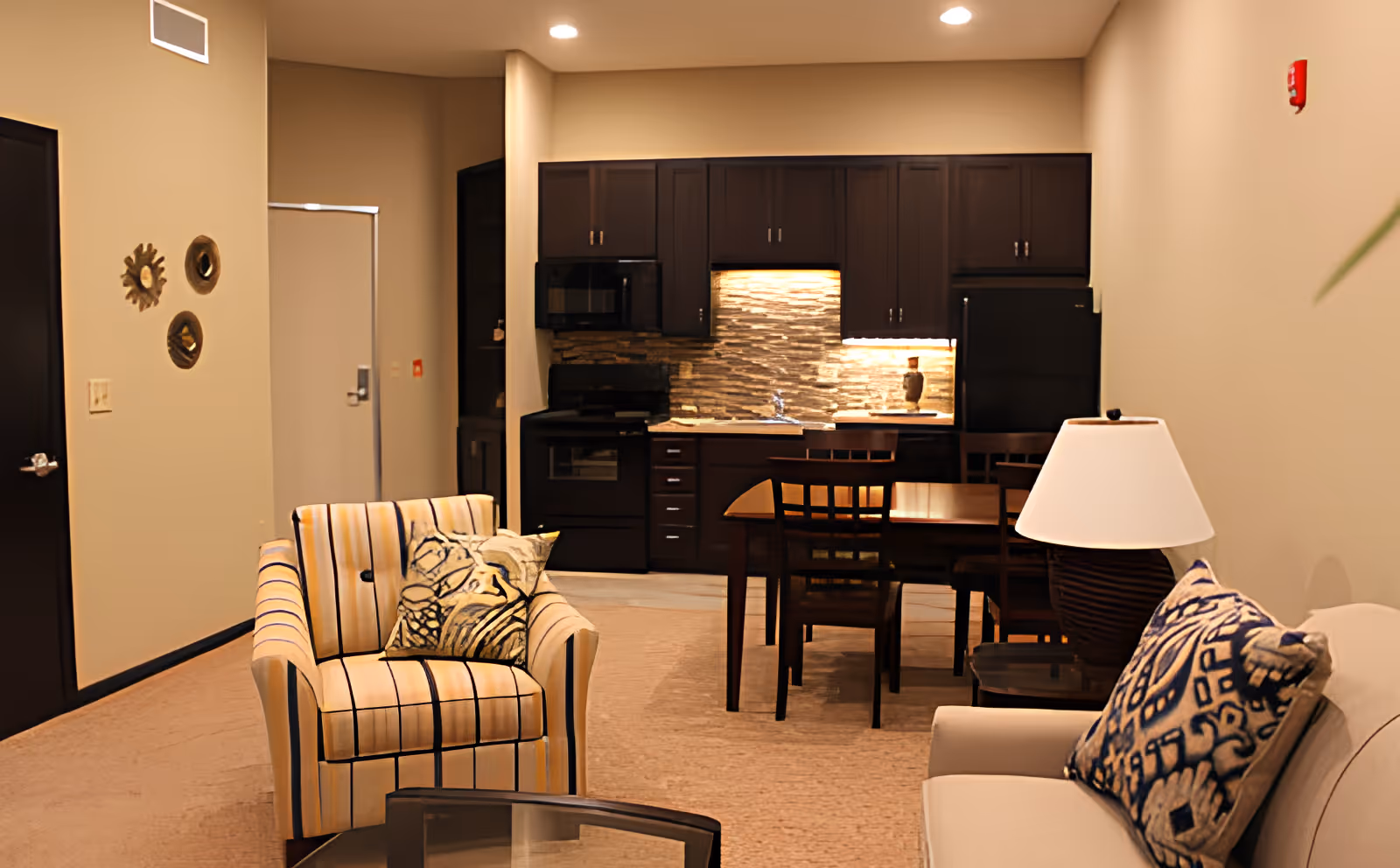 Interior view of a senior living community apartment featuring a cozy living area with a striped armchair and a beige sofa adorned with patterned cushions. A wooden dining table with chairs is positioned near a modern kitchen with dark cabinetry, a tiled backsplash, and under-cabinet lighting. The walls are painted beige, and the room is softly lit with recessed ceiling lights.