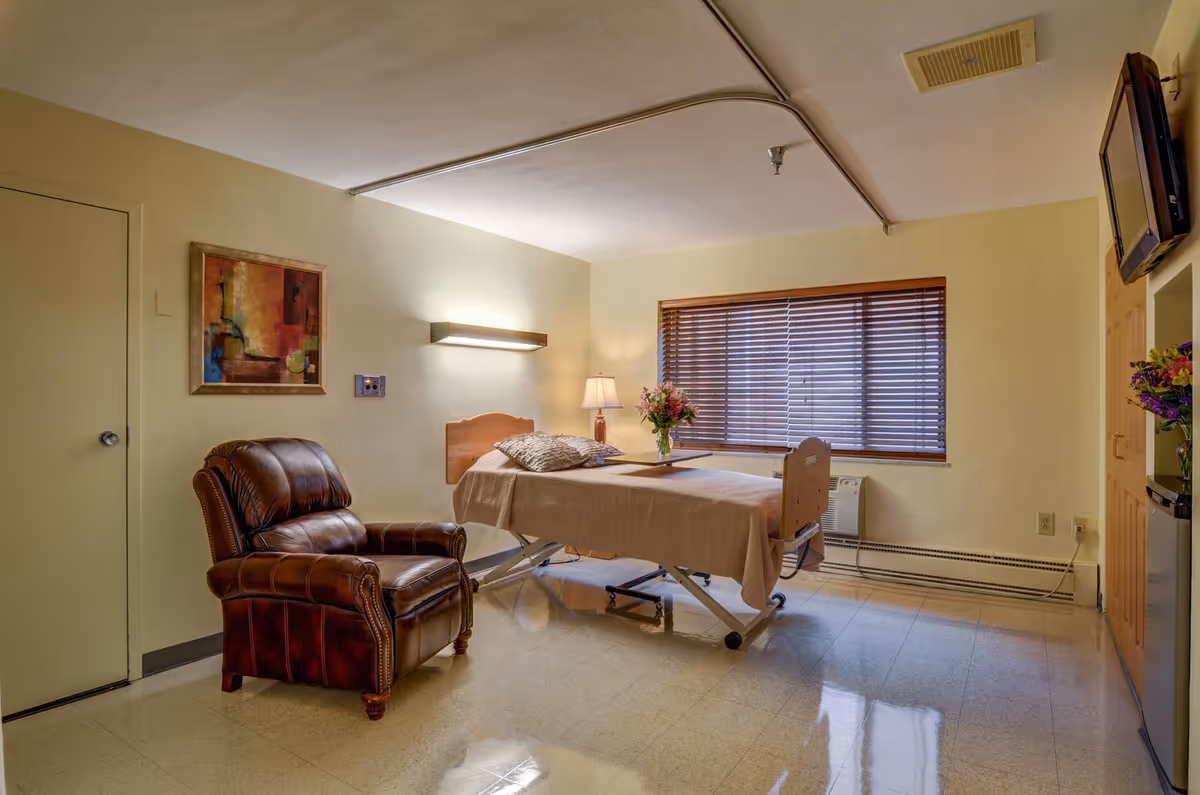A well-lit room in a senior living facility featuring a hospital-style bed with beige bedding and pillows, a brown leather recliner chair, a side table with a lamp and a vase of flowers, a window with wooden blinds, a wall-mounted TV, and a piece of abstract artwork on the wall.