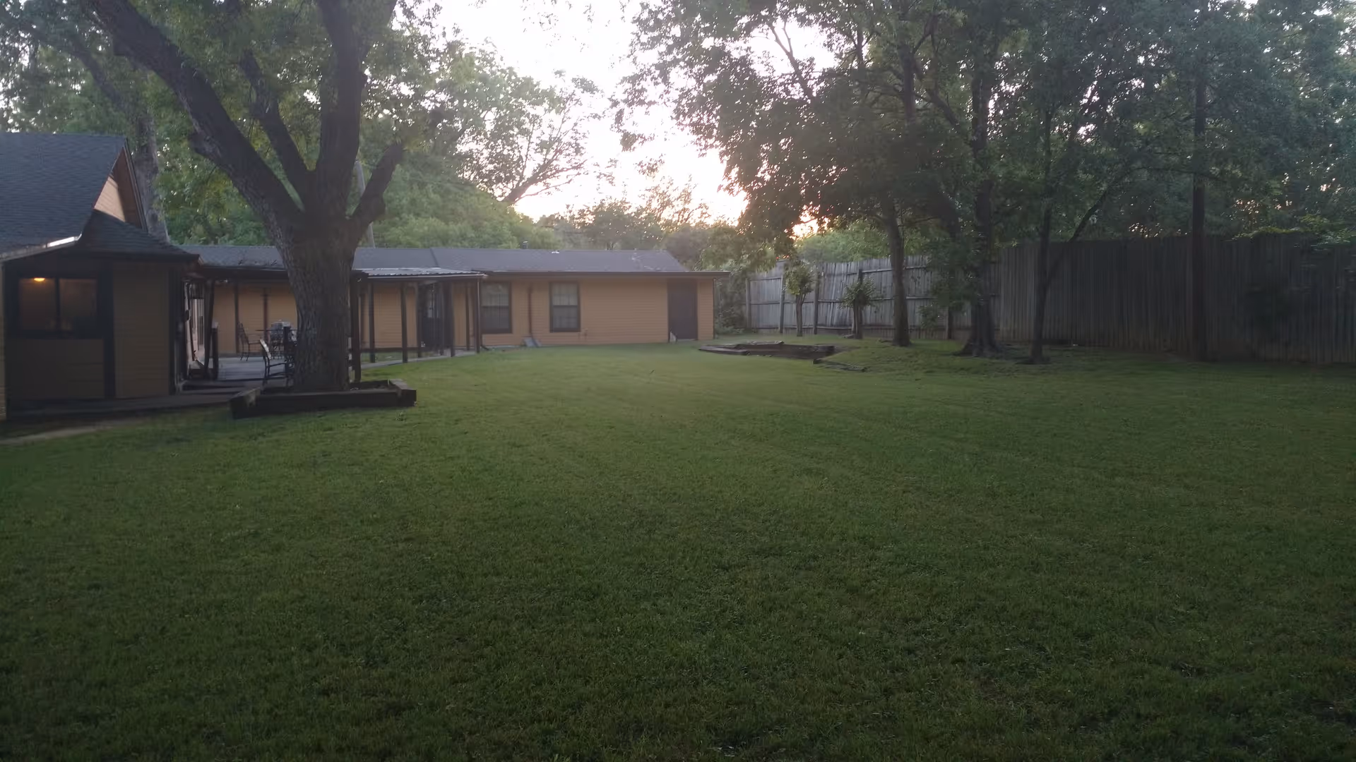 A large grassy backyard area with several mature trees and a wooden fence surrounding the space. There is a single-story building with a covered porch on the left side and another building in the background. The scene is lit by natural light, suggesting early morning or late afternoon.
