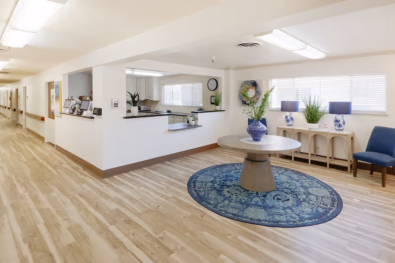 Bright and clean interior space with light wood flooring, a round table with a blue patterned rug underneath, a blue vase with flowers on the table, two blue lamps on a console table against the wall, a blue chair, and a hallway leading to rooms on the left side.