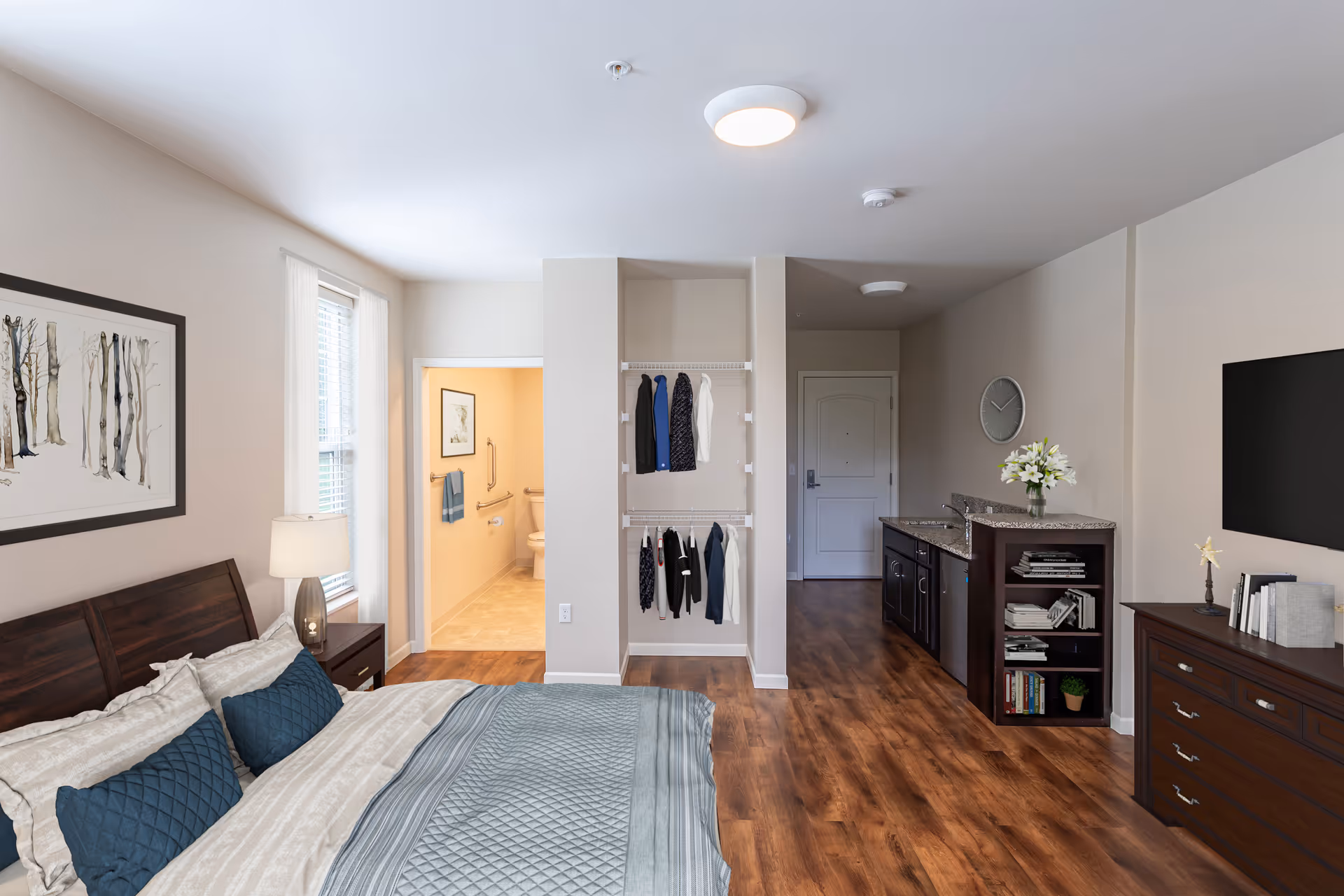A furnished studio-style bedroom with a bed, open closet, kitchenette and a bathroom doorway, all with wood floors and neutral decor.