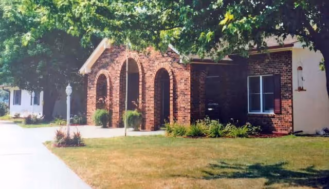 Exterior view of a single-story brick building with arched entryways and a well-maintained lawn with shrubs and trees, under a sunny sky.
