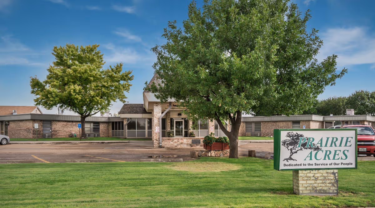Exterior view of Prairie Acres Nursing Home/Friona Heritage Estates building with a green lawn, trees, and a sign in front that reads 'PRAIRIE ACRES Dedicated to the Service of Our People'. The building is a single-story brick structure with a covered entrance.