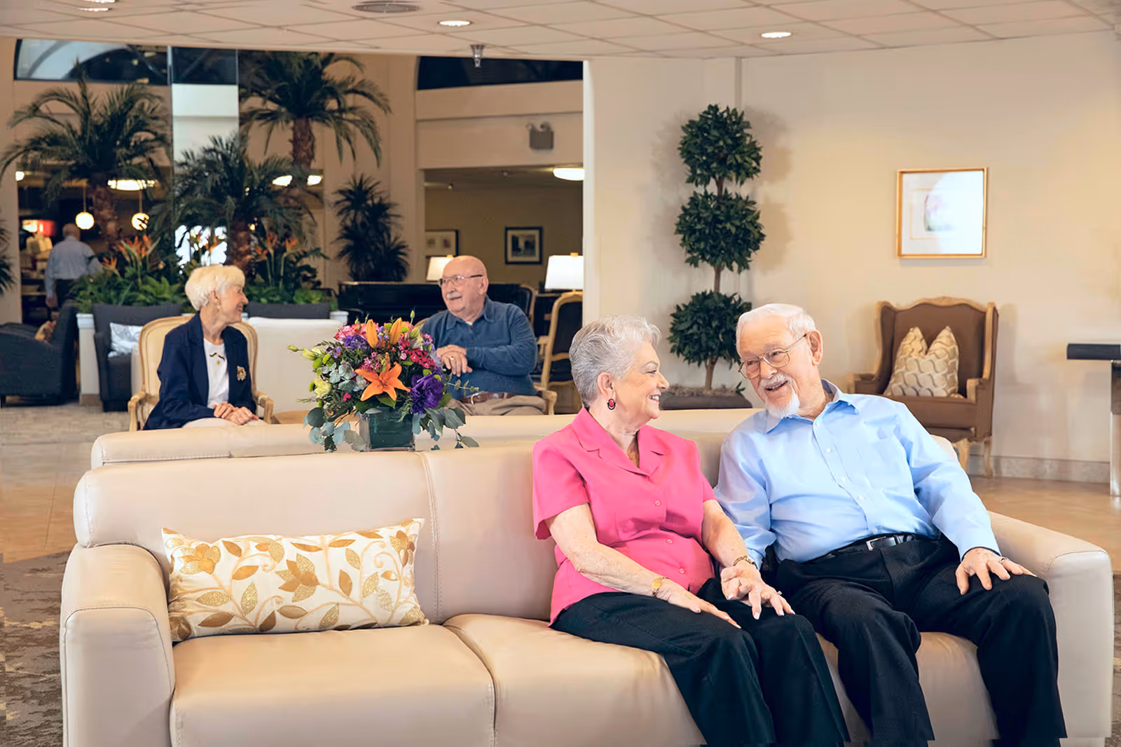 Senior residents sitting and chatting on sofas in a bright lounge area with plants and a floral centerpiece.