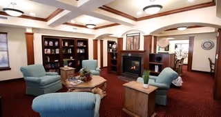 A cozy living room area with four blue upholstered armchairs arranged around a wooden coffee table. The room features a fireplace with built-in shelving on either side, decorative ceiling beams, and warm lighting fixtures. The carpet is red, and there are decorative items on the shelves and tables. An adjacent dining area is visible in the background.