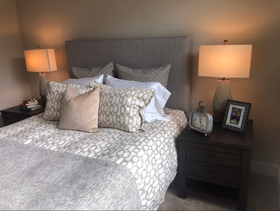 A neatly made bed with a gray upholstered headboard, patterned beige and white bedding, and multiple pillows. On each side of the bed are dark wooden nightstands with table lamps, an alarm clock, a framed photo, and a small stack of books.