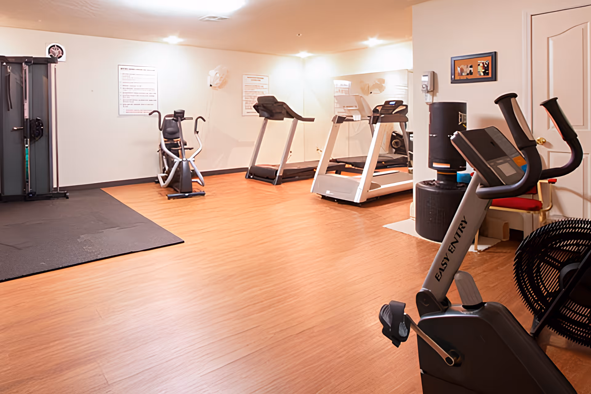 A fitness room with exercise equipment including two treadmills, a stationary bike labeled 'Easy Entry', an elliptical machine, and a multi-function weight machine. The room has wood flooring, white walls, and bright overhead lighting.