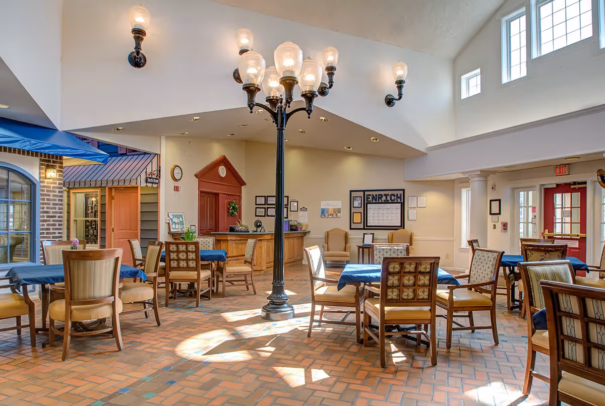 A bright and spacious common area with multiple tables covered in blue tablecloths and surrounded by wooden chairs. The room features a tall black streetlamp-style light fixture in the center, brick-patterned flooring, and high ceilings with multiple windows letting in natural light. There is a reception desk in the background, a bulletin board labeled 'ENRICH,' and a red door on the right side.