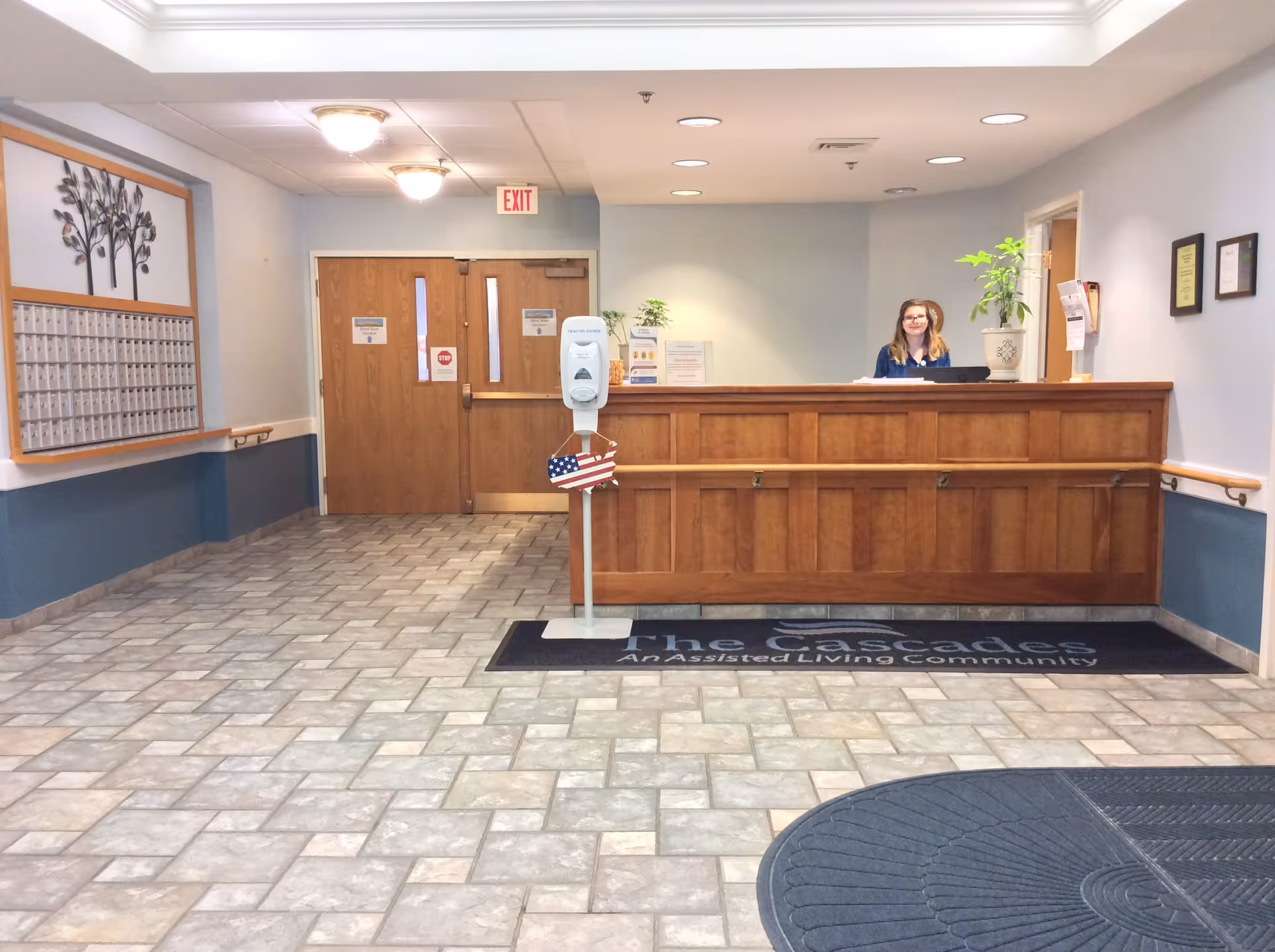 Reception area of The Cascades Assisted Living facility with a wooden front desk, a smiling receptionist behind the desk, a hand sanitizer stand with an American flag decoration, tiled floor, double wooden doors with exit signs, and a wall-mounted mailbox unit with a decorative tree above it.