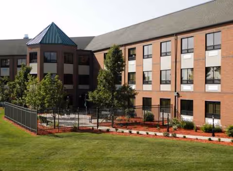 Brick senior living facility exterior with a fenced courtyard and manicured lawn.