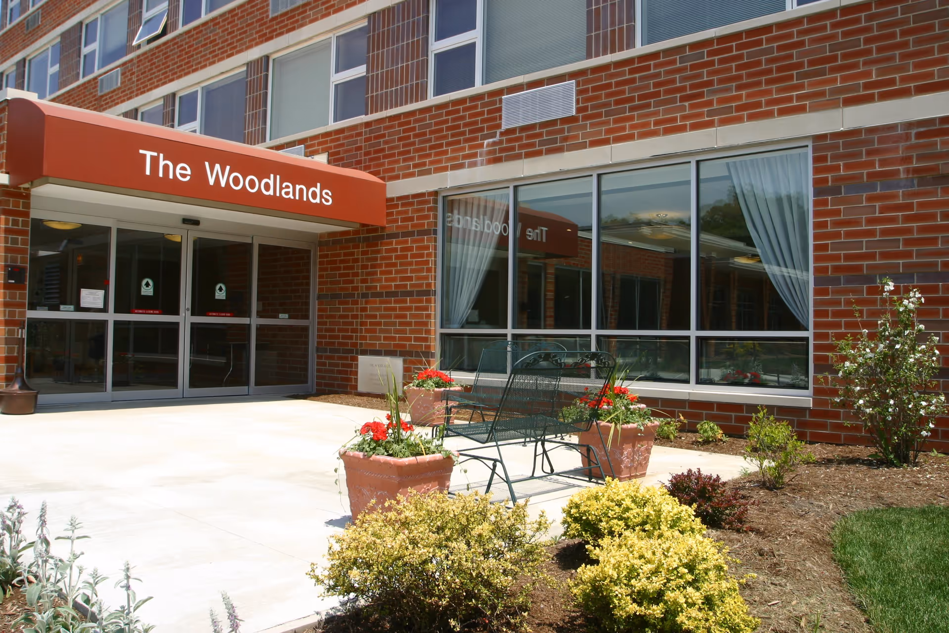 Entrance to a brick building with a red awning that reads 'The Woodlands'. There are large windows with white curtains and a small outdoor seating area with a metal bench surrounded by flower pots and landscaping.
