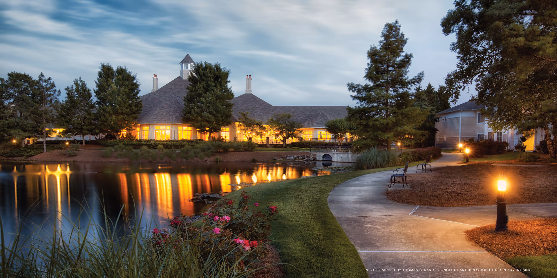 Dusk view of a lit senior living building by a reflective pond with a walking path, benches, and lamps.