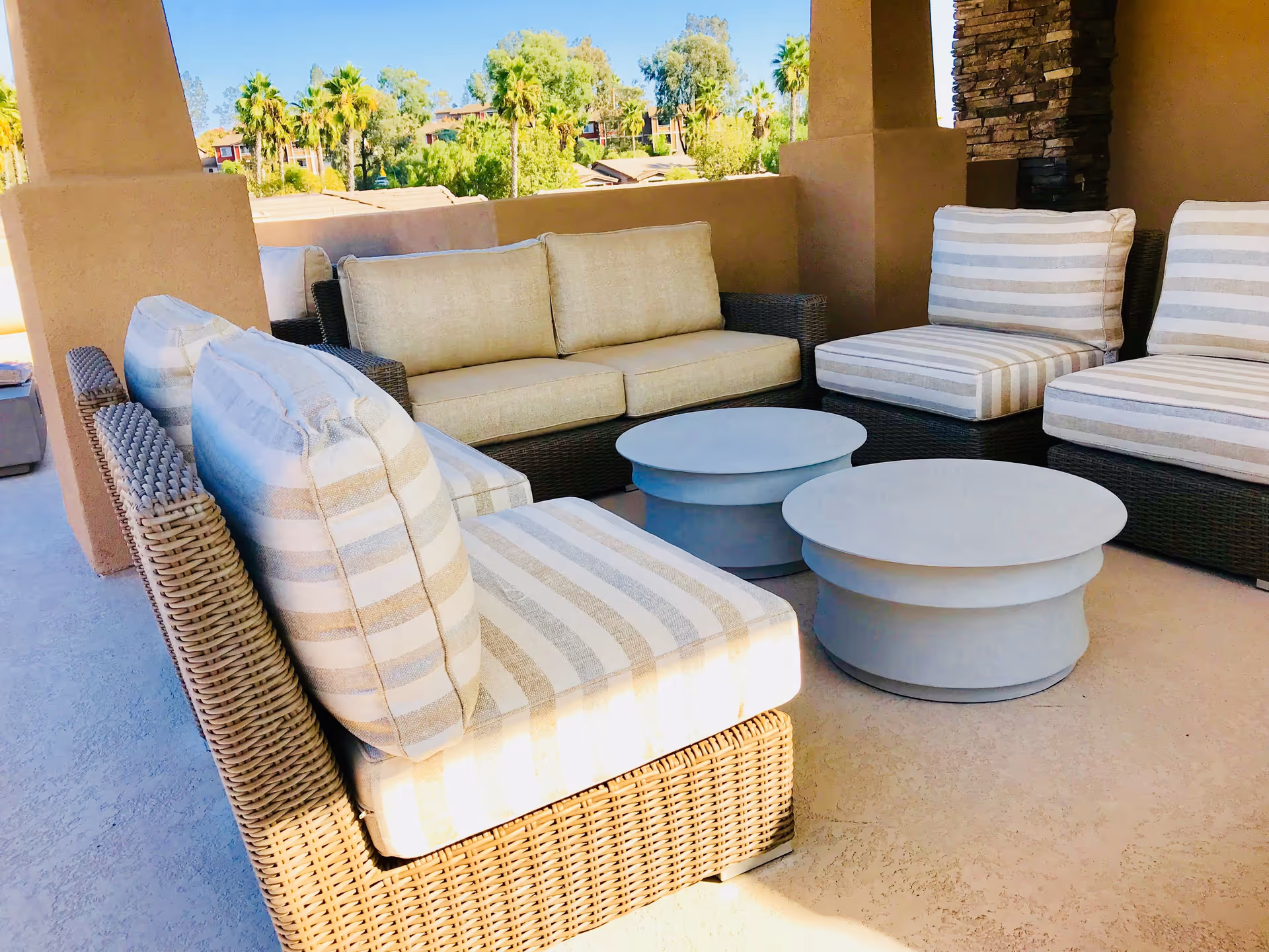Covered outdoor patio with wicker seating, striped cushions, and round coffee tables overlooking palm trees.