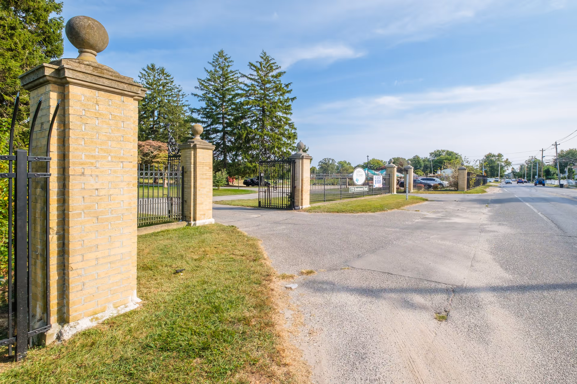 Entrance gate with brick pillars and black wrought iron fencing leading into a green area with trees and parked cars, adjacent to a paved road with vehicles and utility poles under a partly cloudy sky.