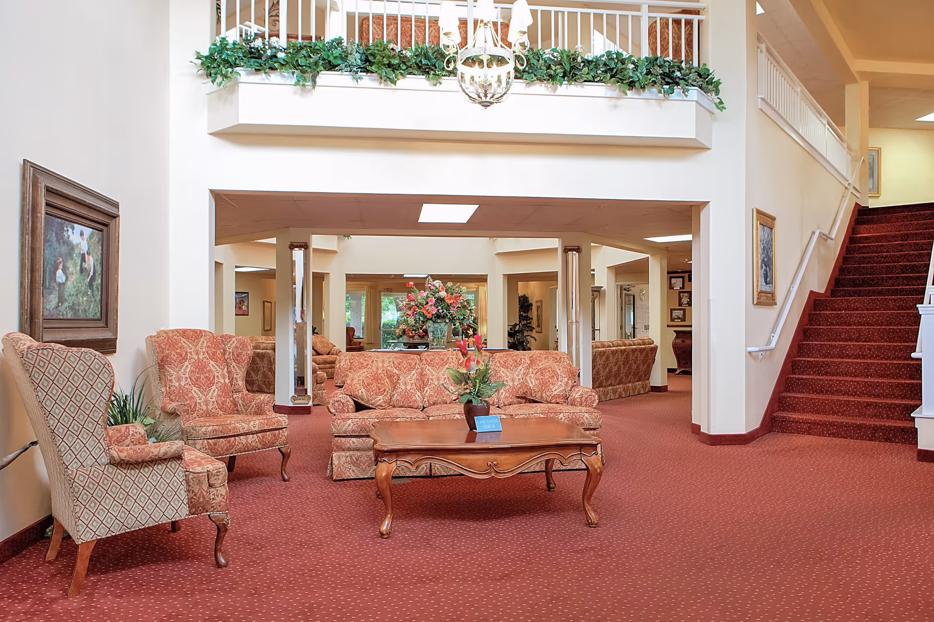 A spacious living room area with red patterned carpet, a floral upholstered sofa, two matching armchairs, and a wooden coffee table with a flower arrangement. The room has white walls, a staircase with red carpet on the right, and a balcony with green plants hanging over the railing above. There are framed paintings on the walls and a chandelier hanging from the ceiling.